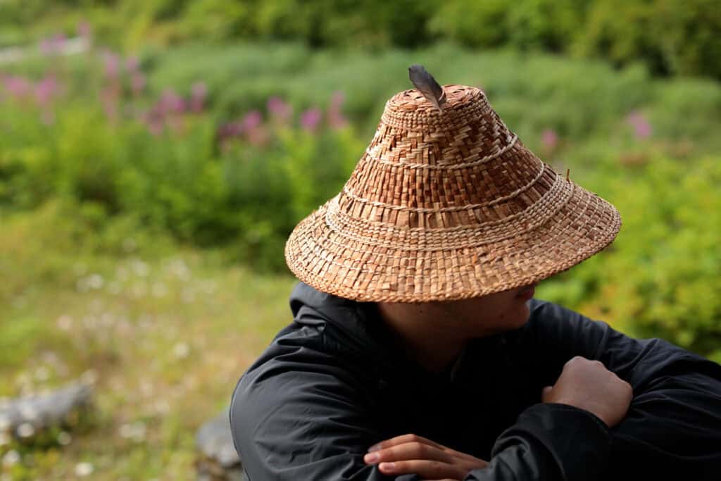 A person in a cedar hat sits in the foreground. Blooming fireweed can be seen in the background.