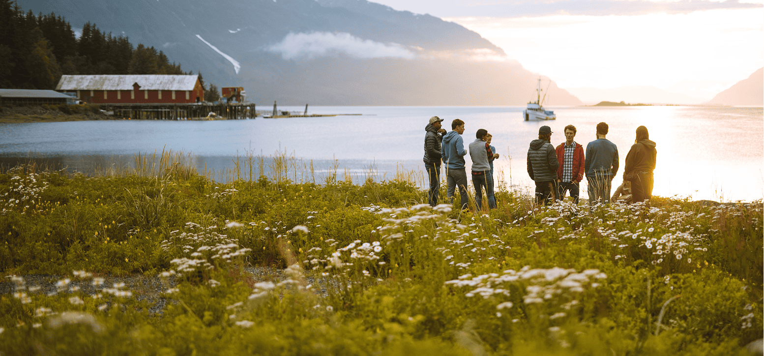 Scenic view of a coastline in Southeast AK with a gathering of people around a campfire in the midground, and blooming plants in the foreground.
