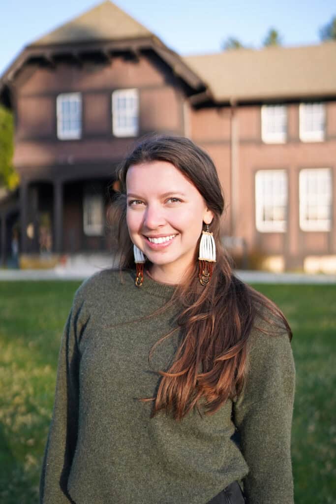 A woman in a green sweater and black pants standing in front of a house.