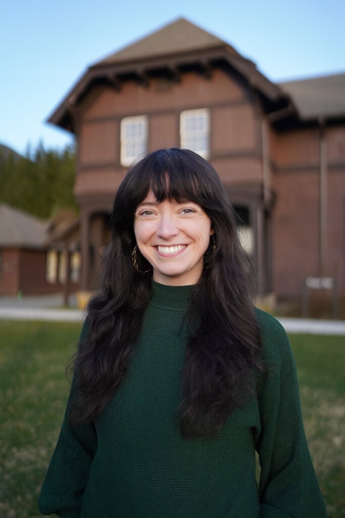 A woman with long black hair and green sweater standing in front of a house.