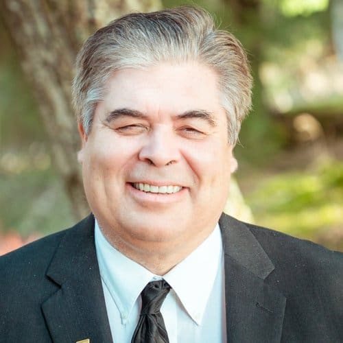 A man in a suit and tie smiling in front of trees.