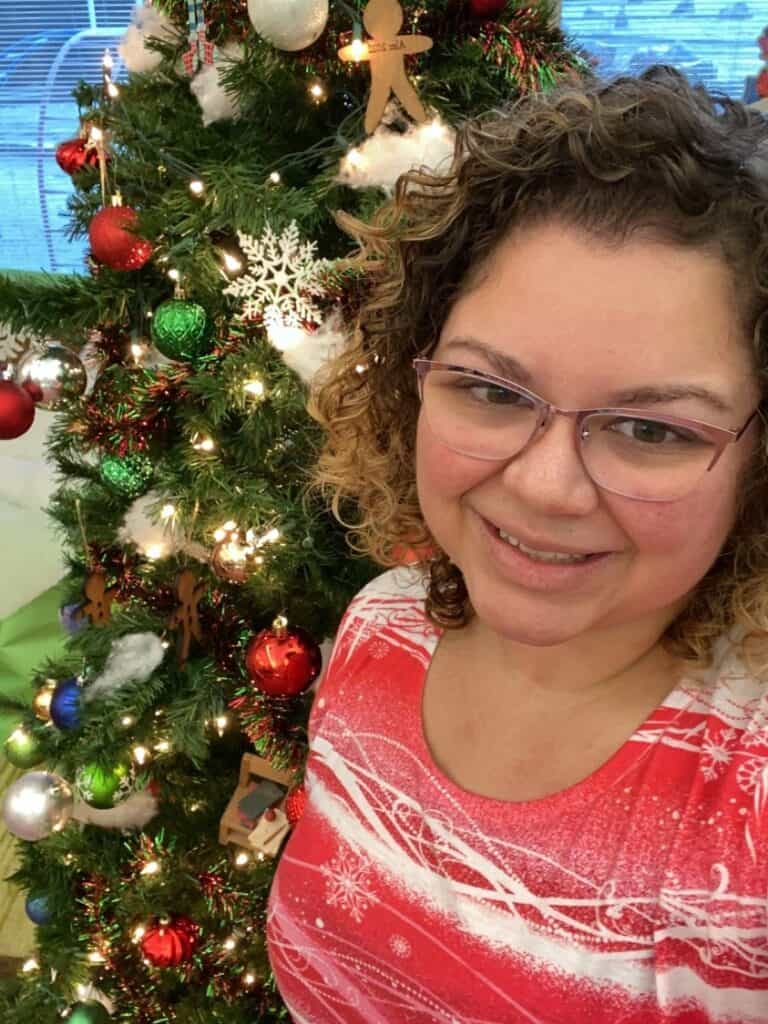 A woman with glasses and a red shirt is standing in front of a christmas tree.