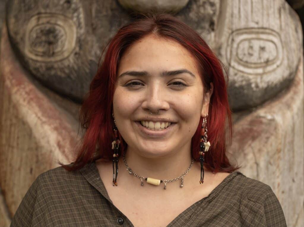 A woman with red hair and earrings standing in front of a totem pole.