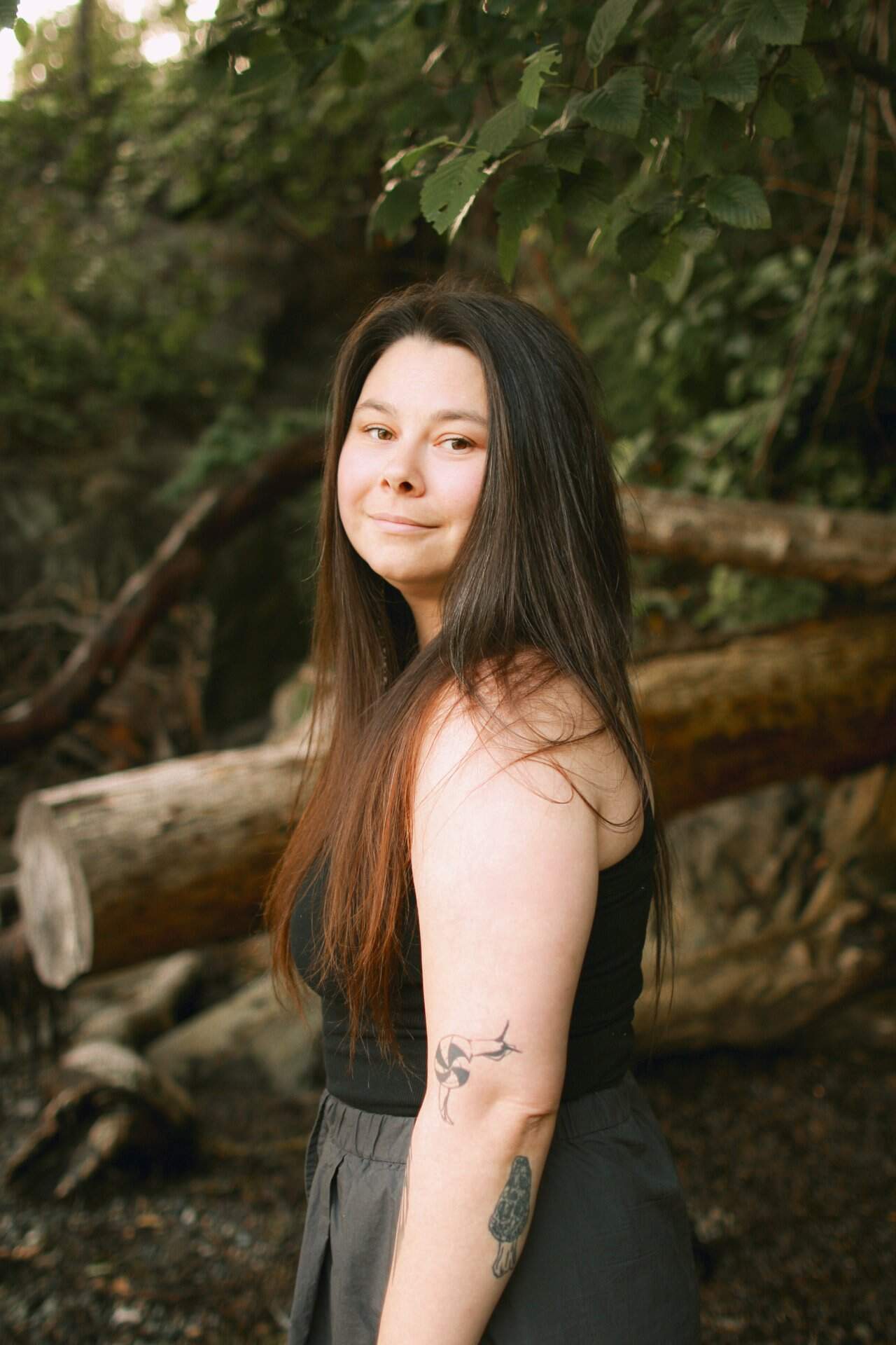 A woman with long hair and tattoos standing in front of a tree.