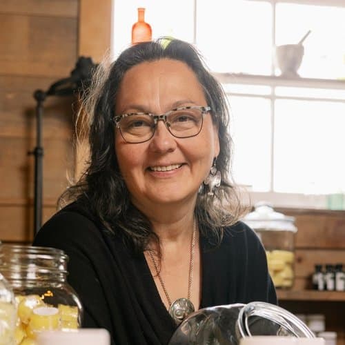 A woman with glasses and a black shirt sitting at a table with jars of botanical products.