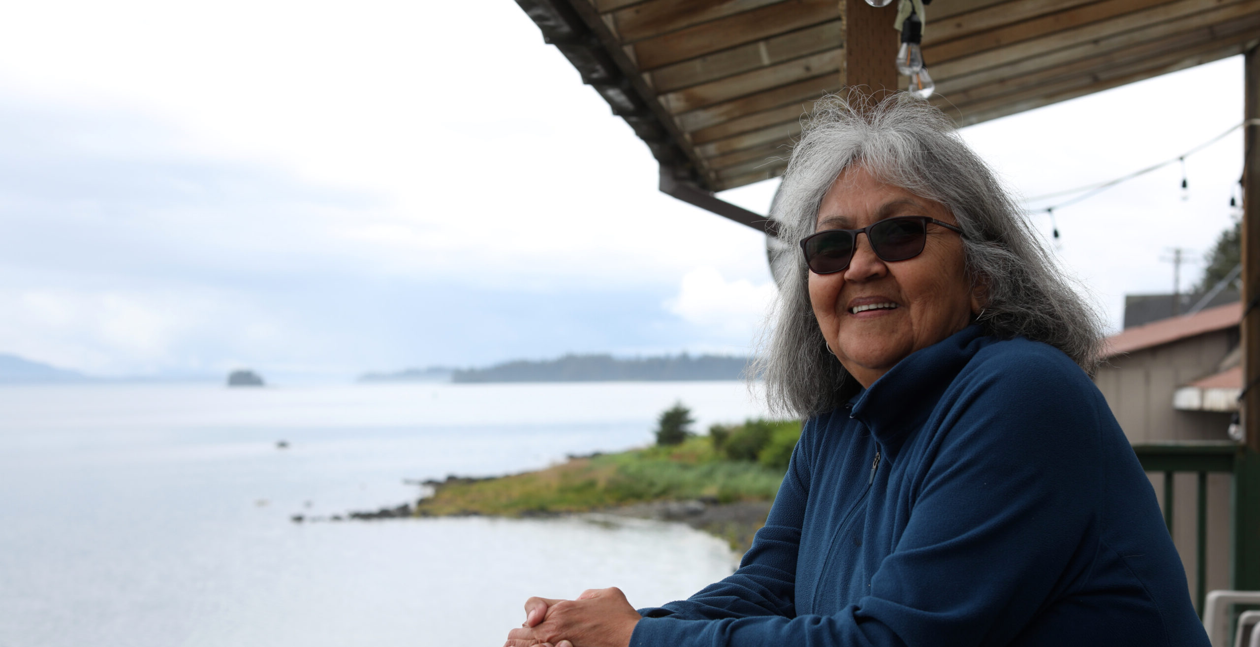 An older woman in sunglasses standing on a deck overlooking the water.