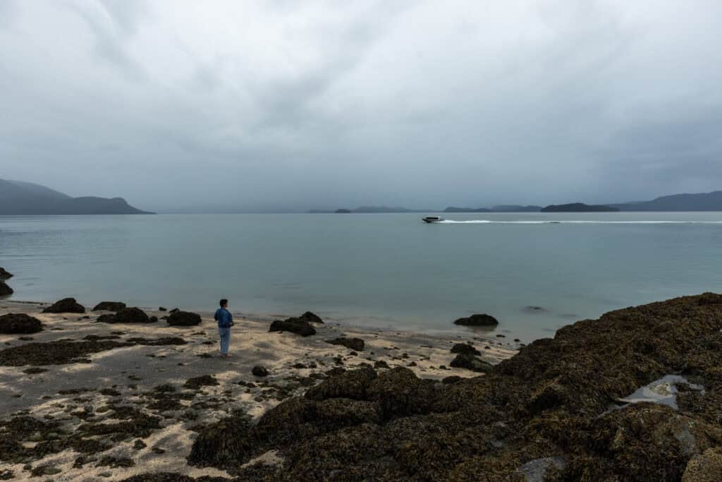 A person stands on the shores of a coast, while a boat speeds by.