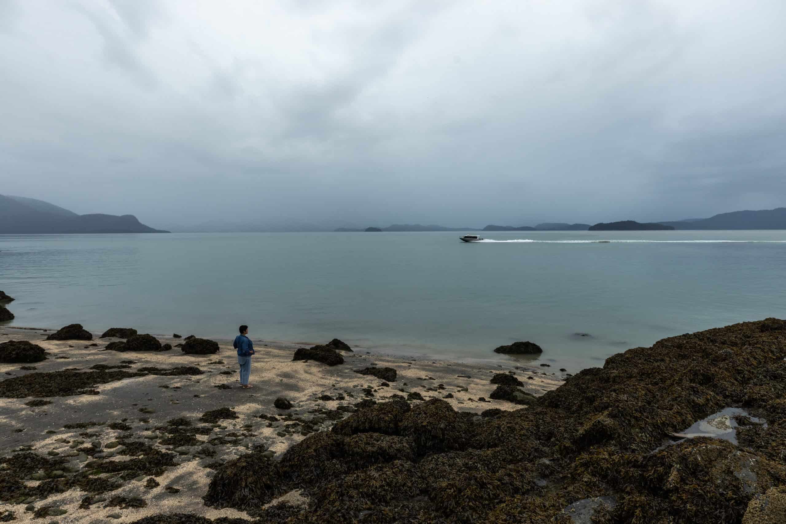 A person stands on the shores of a coast, while a boat speeds by.